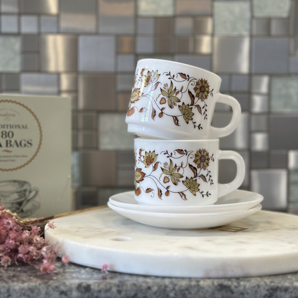 Two white ceramic cups with floral patterns stacked on a saucer, placed on a marble cutting board. A box of tea bags is in the background, and soft pink flowers are in the corner. The setting has a tiled backdrop and a granite counter.