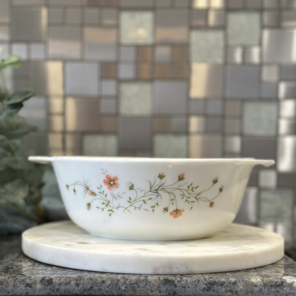 A white bowl with floral designs sits on a marble tray against a tiled backsplash. The design features small, delicate orange flowers and green leaves.