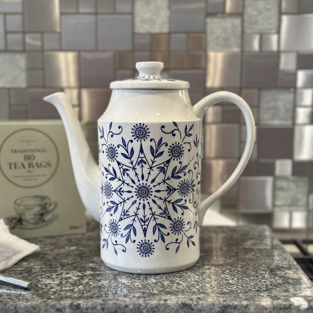 A white ceramic teapot with blue floral patterns sits on a granite countertop. In the background, there's a box of 80 tea bags and a textured tile backsplash in various shades of gray.