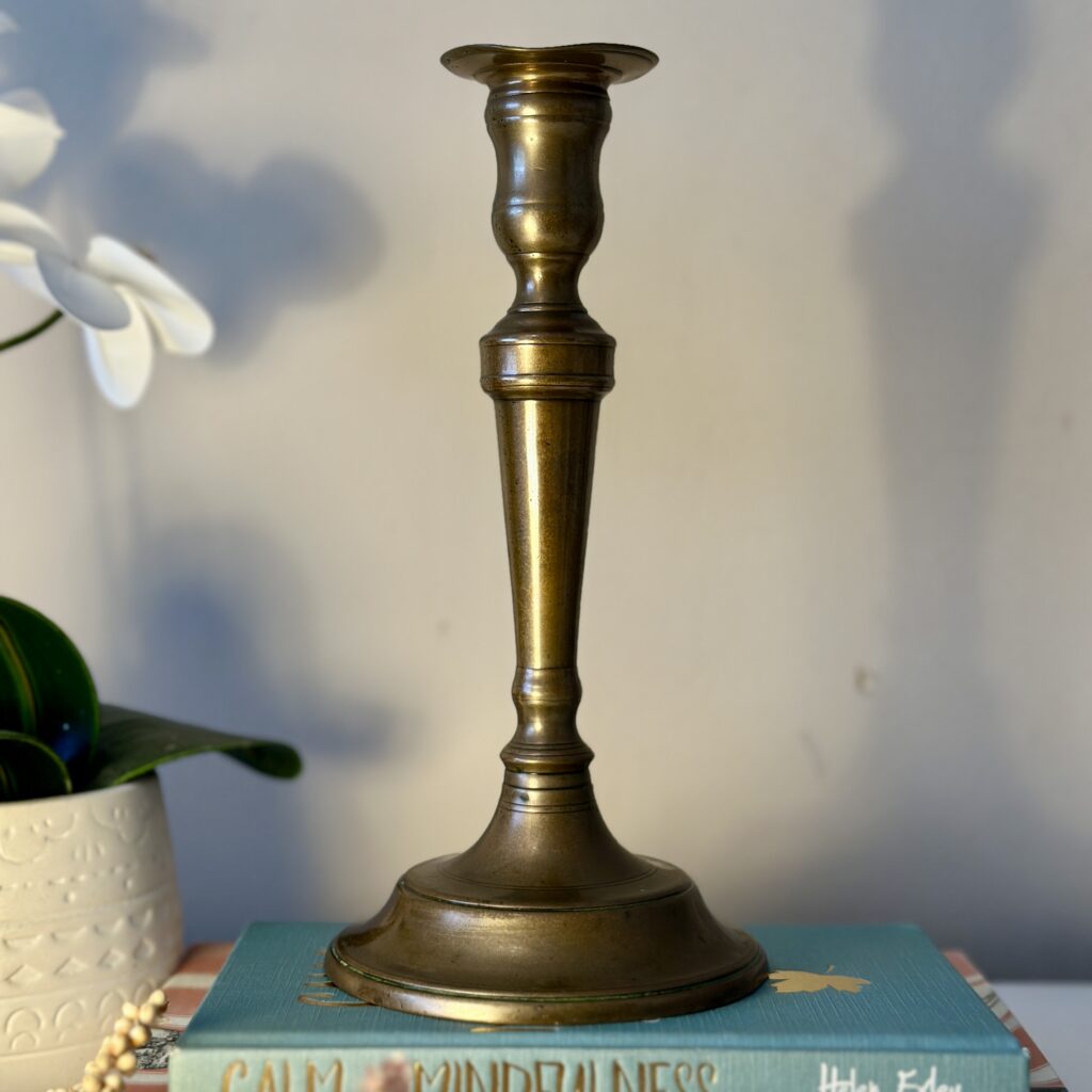 A brass candlestick stands on a stack of books titled "Calm Mindfulness." A potted plant with white flowers is partially visible on the left, against a light-colored wall.